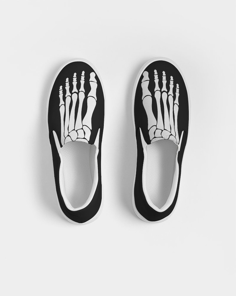 May include: A pair of black canvas slip-on shoes with a white skeleton foot design on the front.