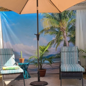 May include: Two striped patio chairs with a tropical beach scene backdrop. A brown umbrella with a white canopy provides shade. A small potted plant sits in front of the chairs.