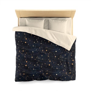 May include: A navy blue duvet cover with a constellation pattern featuring gold and orange stars and lines. The duvet cover is shown on a bed with a white headboard and two white pillows.