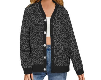 May include: A button-down cardigan with a black and gray leopard print. The cardigan has a V-neck, black trim, and large silver buttons. The sleeves are long, and the fabric appears to be a soft knit. The garment is worn over a white top and blue jeans.
