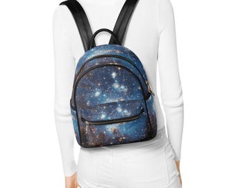 May include: A small backpack featuring a vibrant galaxy print with deep blue, purple, and black hues, accented by numerous bright white stars and nebulae. The bag includes black shoulder straps, a top handle, and a front zippered pocket.