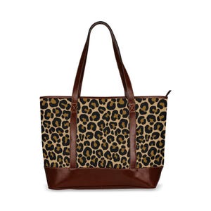 May include: A tote bag featuring a leopard print design in brown and black. It has brown leather handles and a brown leather base. The bag is equipped with a zip closure.