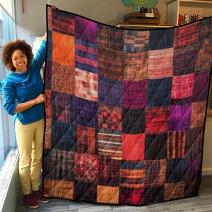 May include: A patchwork quilt with a variety of colors and patterns, including red, orange, brown, purple, and blue. The quilt is made of squares and rectangles, and the seams are visible. The quilt is folded in half and is lying on a wooden floor.