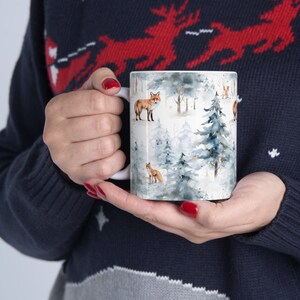 K&ouml;nnte beinhalten: Wei&szlig;e Keramik-Tasse mit einem Winterwald-Design, das Kiefern und F&uuml;chse im Aquarellstil in Blau-, Grau- und Braunt&ouml;nen zeigt. Die Tasse hat einen wei&szlig;en Henkel. Die Tasse wird von einer Person gehalten, die einen marineblauen Pullover tr&auml;gt.