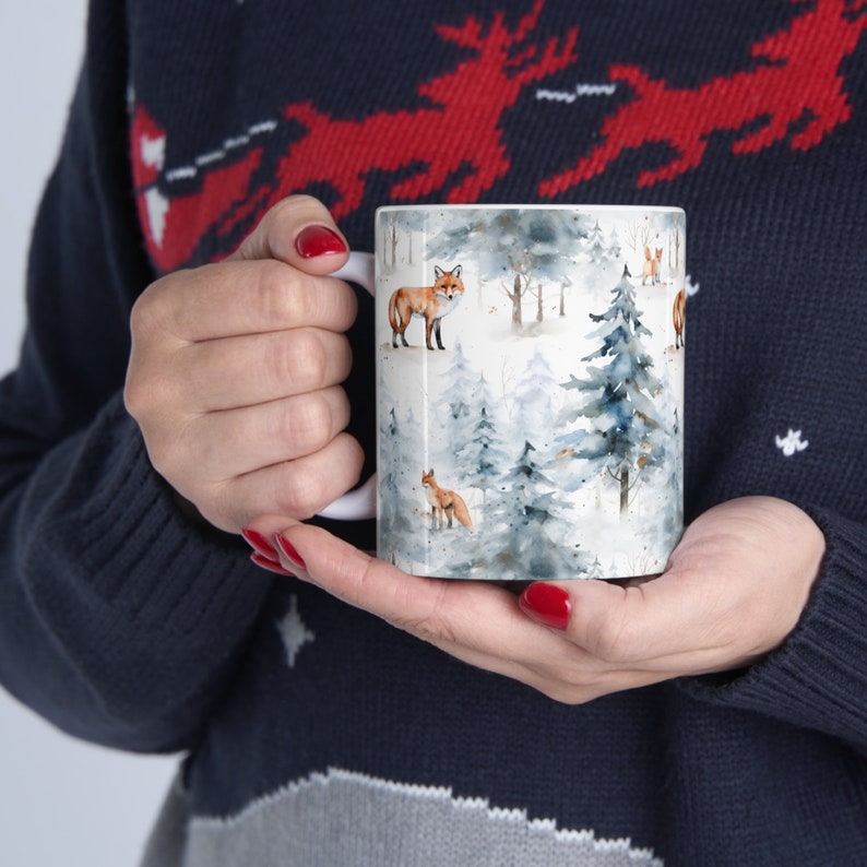 K&ouml;nnte beinhalten: Wei&szlig;e Keramik-Tasse mit einem Winterwald-Design, das Kiefern und F&uuml;chse im Aquarellstil in Blau-, Grau- und Braunt&ouml;nen zeigt. Die Tasse hat einen wei&szlig;en Henkel. Die Tasse wird von einer Person gehalten, die einen marineblauen Pullover tr&auml;gt.