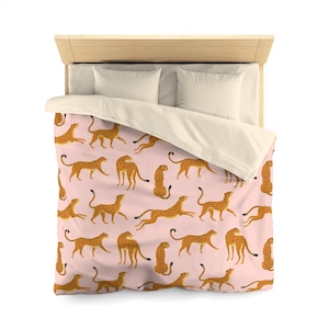 Könnte beinhalten: Ein rosafarbener Bettbezug mit einem sich wiederholenden Muster aus orangefarbenen und braunen Geparden. Der Bettbezug liegt auf einem Bett mit zwei weißen Kissen.