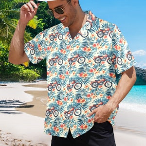Puede incluir: Una camisa de manga corta con botones y estampado tropical con motocicletas antiguas y hojas de palmera. La camisa es de color crema con detalles en rojo, azul y verde.