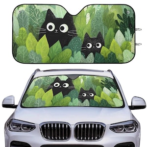 May include: Car windshield sunshade featuring a whimsical design of three black cats peeking out from lush green foliage. The cats have large, expressive eyes. The sunshade is designed to fit most car windshields.
