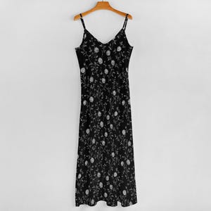 May include: A black midi dress with a V-neck and spaghetti straps, adorned with a white floral pattern. The dress hangs on a wooden hanger against a white background, showcasing a delicate and feminine design.