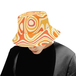 Puede incluir: Un sombrero de pescador con un diseño retro psicodélico. El sombrero presenta un patrón arremolinado en tonos naranja, amarillo y crema. El borde está bordeado con costuras negras. El sombrero lo lleva una persona que lleva una camiseta negra.