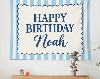 May include: A rectangular fabric wall hanging featuring a light blue and white vertical striped background. A large, central cream-colored scalloped rectangle displays the dark blue text "HAPPY BIRTHDAY Noah" in a mix of block and script fonts.