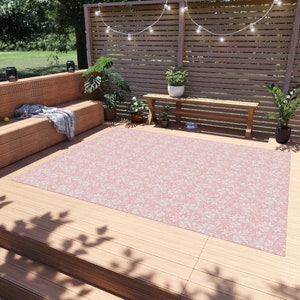 May include: A pink outdoor rug with a white floral pattern. The rug is laid out on a wooden deck with a wooden bench and plants in the background.