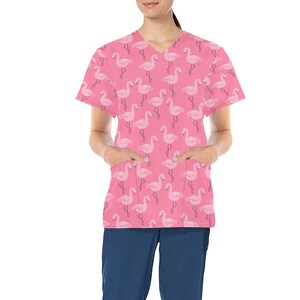 Puede incluir: Camiseta de trabajo rosa con estampado de flamencos. La camiseta tiene cuello en V y mangas cortas. Tiene dos bolsillos en la parte delantera.