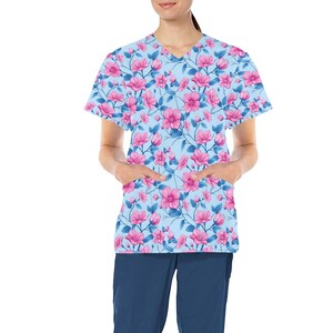 Puede incluir: Una blusa azul claro con cuello en V con un estampado floral de flores rosas y hojas azules. La blusa tiene dos bolsillos delanteros y mangas cortas. También se ve un pantalón azul marino.