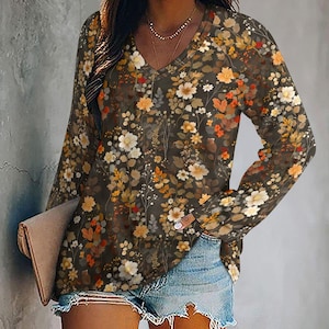 Può includere: Un top a maniche lunghe con scollo a V con un motivo floreale nei toni del marrone, arancione e bianco. Il top è abbinato a pantaloncini di jeans. Il motivo floreale copre l'intero top, creando un look elegante.