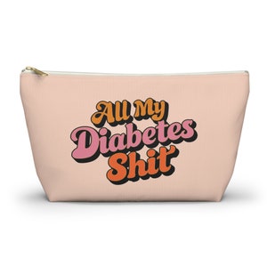 Puede incluir: Una bolsa de maquillaje de lona rosa con cierre de cremallera. La bolsa tiene un gráfico de estilo retro que dice "All My Diabetes Shit" en letras naranja, rosa y negras.