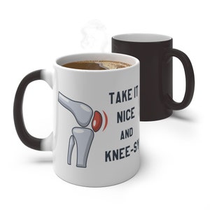 May include: White coffee mug with a black handle. The mug has a black and white image of a knee joint with a red spot on it. The text "TAKE IT NICE AND  KNEE-SY" is printed on the mug.