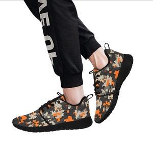 Puede incluir: Un par de zapatillas negras con un patrón de camuflaje en naranja, gris y negro. Las zapatillas tienen una suela blanca y un cierre de cordones.