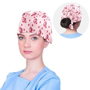 Op de afbeelding: Een roze en rode gepatroneerde chirurgische scrub-muts met een strik aan de achterkant. De muts heeft een ontwerp van rode kersen en witte stippen.