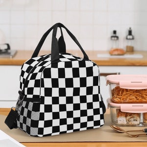 Puede incluir: Bolsa de almuerzo a cuadros en blanco y negro con asa negra y bolsillo de malla en el lateral. La bolsa está sobre una mesa de madera.