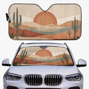 Op de afbeelding: Auto zonnescherm met een woestijnlandschap ontwerp. Het heeft een grote oranje zon, cactussen en gelaagde bergen in tinten oranje, bruin en blauw. Het zonnescherm wordt opgevouwen en in een auto getoond.