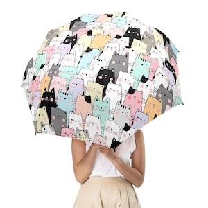 Puede incluir: Un paraguas colorido con un patrón repetido de gatos de dibujos animados en varios colores. Los gatos tienen caras sonrientes y están sobre un fondo blanco. El paraguas es sostenido por una persona.