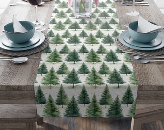 Puede incluir: Un camino de mesa rectangular con un patrón repetido de árboles de Navidad verdes en acuarela sobre un fondo beige claro. El camino de mesa está colocado sobre una mesa de madera, con cubiertos, copas de vino y un jarrón con lirios rosas.