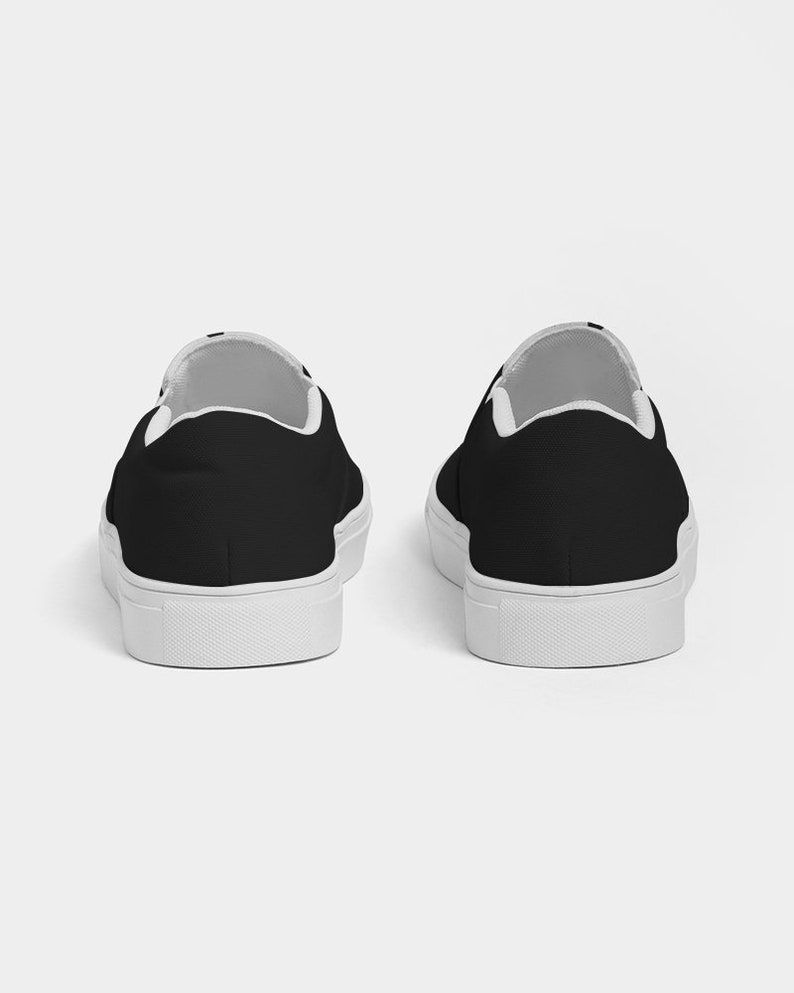 May include: A pair of black canvas slip-on shoes with white soles. The shoes are shown from the back.