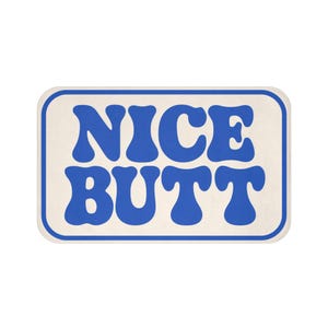 May include: A rectangular bath mat with rounded corners featuring a cream-colored background and a thick blue border. The text NICE BUTT is printed in the center in a bold, wavy, blue sans-serif font that fills the majority of the mat surface.