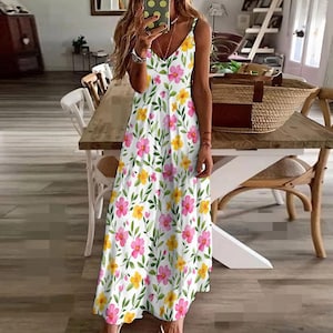 Puede incluir: Un vestido maxi blanco con estampado floral con flores rosas y amarillas. El vestido tiene un escote en V y tirantes finos.