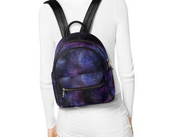 May include: A small backpack featuring a vibrant galaxy print with deep purple, blue, and black hues, accented by white star-like speckles. The bag includes black shoulder straps, a top handle, and a front zippered pocket with gold-toned hardware.