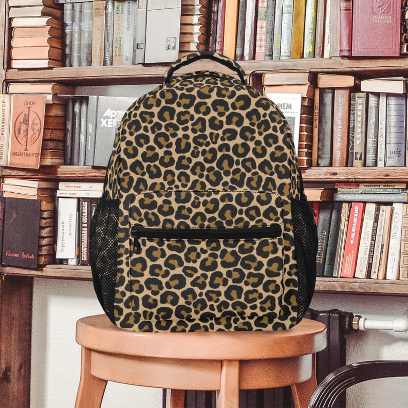 May include: A brown and black leopard print backpack with a black zipper and mesh side pockets. The backpack is sitting on a wooden stool.