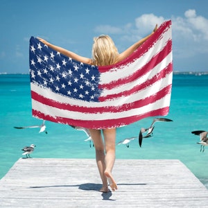 Peut inclure: Une femme portant une serviette de plage avec un motif de drapeau américain délavé. La serviette est rouge, blanche et bleue. La femme est debout sur une jetée en bois avec l'océan en arrière-plan.