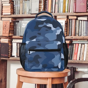 Puede incluir: Una mochila azul de camuflaje con una cremallera negra y bolsillos laterales de malla. La mochila está sentada en un taburete de madera.