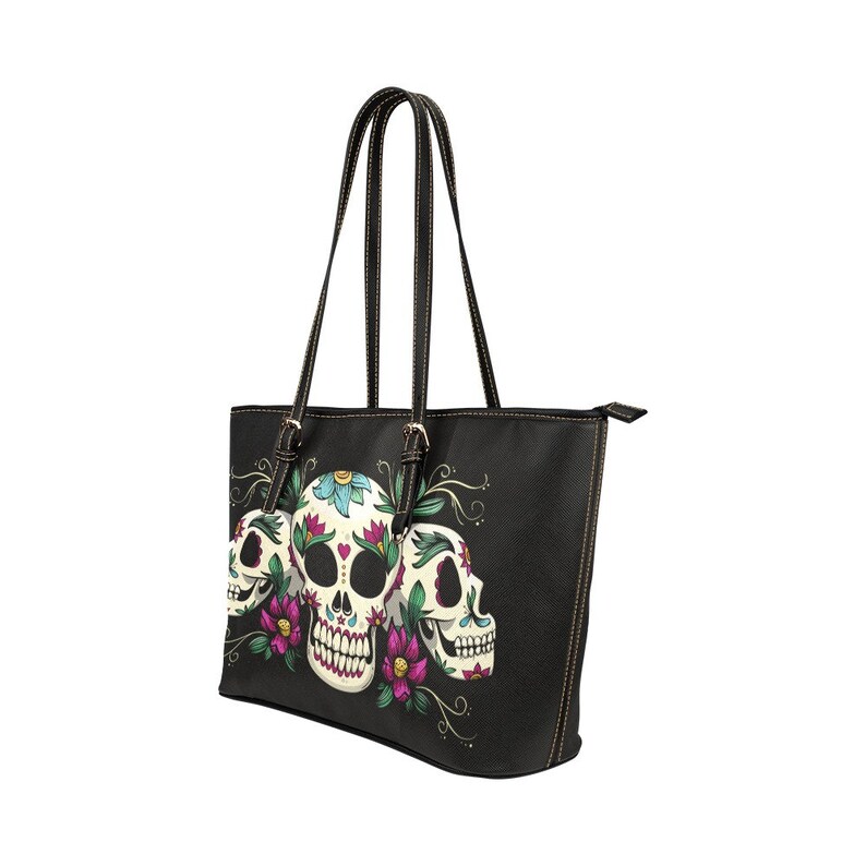 May include: Black tote bag with a design of three sugar skulls with floral accents. The skulls are white with colorful flowers and details.