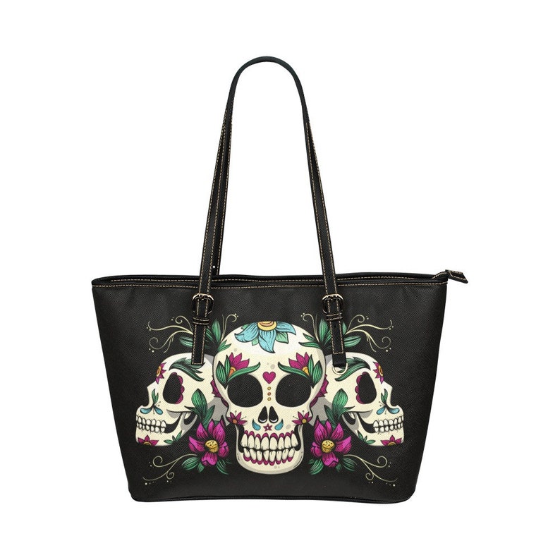 May include: Black tote bag with a design of three sugar skulls with floral accents. The skulls are white with black eyes and pink, blue, and green flowers.