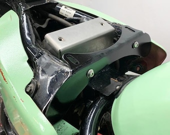 May include: Close-up of a motorcycle's rear fender and frame. The fender is painted a light green color and the frame is black. A silver metal box is mounted to the frame, and a black metal bracket is attached to the fender.