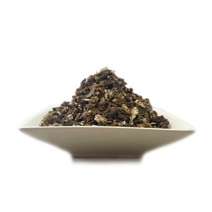 May include: A mound of dried, dark brown, and tan root pieces piled high in a shallow, square, off-white ceramic bowl. The texture of the roots is rough, fibrous, and irregular, with varying shades of brown and small flecks of lighter material.