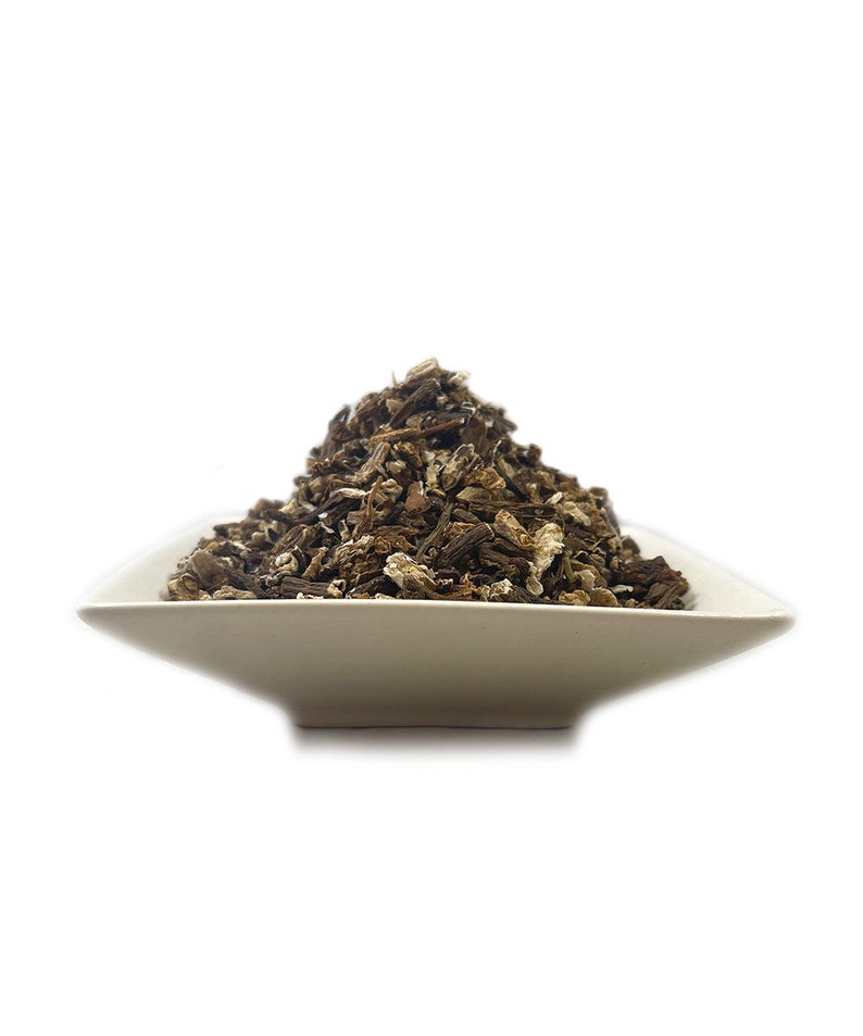 May include: A mound of dried, dark brown, and tan root pieces piled high in a shallow, square, off-white ceramic bowl. The texture of the roots is rough, fibrous, and irregular, with varying shades of brown and small flecks of lighter material.