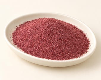 May include: A mound of fine, deep magenta-colored powder is contained within a smooth, oval-shaped white ceramic dish. The texture of the powder appears granular and uniform, filling the center of the shallow bowl with a vibrant, dark red hue.