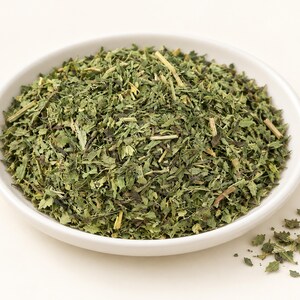 May include: A white ceramic bowl filled with a mound of dried, shredded green herbs. The mixture consists of small, jagged leaf fragments and thin, light-colored woody stems. A few loose pieces of the dried green herb are scattered outside the bowl.