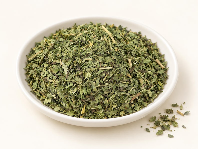 May include: A white ceramic bowl filled with a mound of dried, shredded green herbs. The mixture consists of small, jagged leaf fragments and thin, light-colored woody stems. A few loose pieces of the dried green herb are scattered outside the bowl.