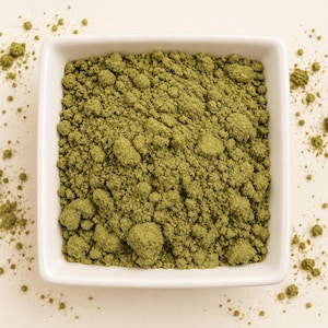 May include: A square white ceramic bowl filled with a fine, textured olive green powder. The powder has a granular consistency with small clumps throughout, and a small amount of the green substance is scattered outside the bowl on the surface.