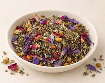 May include: A white bowl filled with a loose-leaf herbal tea blend containing dried green herbs, vibrant pink rose petals, bright blue cornflower petals, and small yellow chamomile flowers, with a few loose pieces scattered around the base.