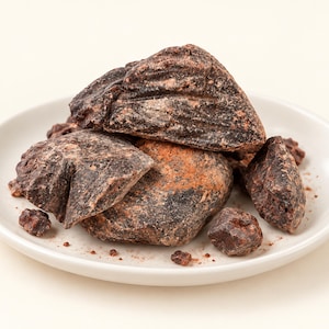 May include: A pile of dark brown, irregularly shaped chunks of raw cacao butter or chocolate mass resting on a small, round, white ceramic plate. The pieces have a rough, textured surface with visible variations in color, ranging from deep brown to reddish-brown tones, with small crumbs scattered on the plate.