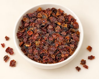 May include: A white bowl filled with dried, crushed rose hips. The pieces are irregular in shape and range in color from deep burgundy and dark red to brownish-orange. Several individual dried rose hip fragments are scattered outside the bowl.