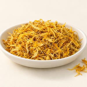 May include: A white ceramic bowl filled with a mound of dried, shredded yellow and orange flower petals. The petals are thin, curled, and textured, creating a dense pile within the bowl, with a few loose petals scattered near the base.
