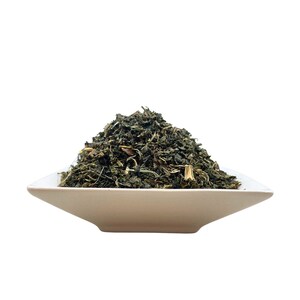 May include: A mound of dried, dark green herbal leaves and small stems piled high in a square, off-white ceramic bowl. The texture of the dried plant matter is coarse and varied, featuring shades of deep forest green and lighter brown accents.