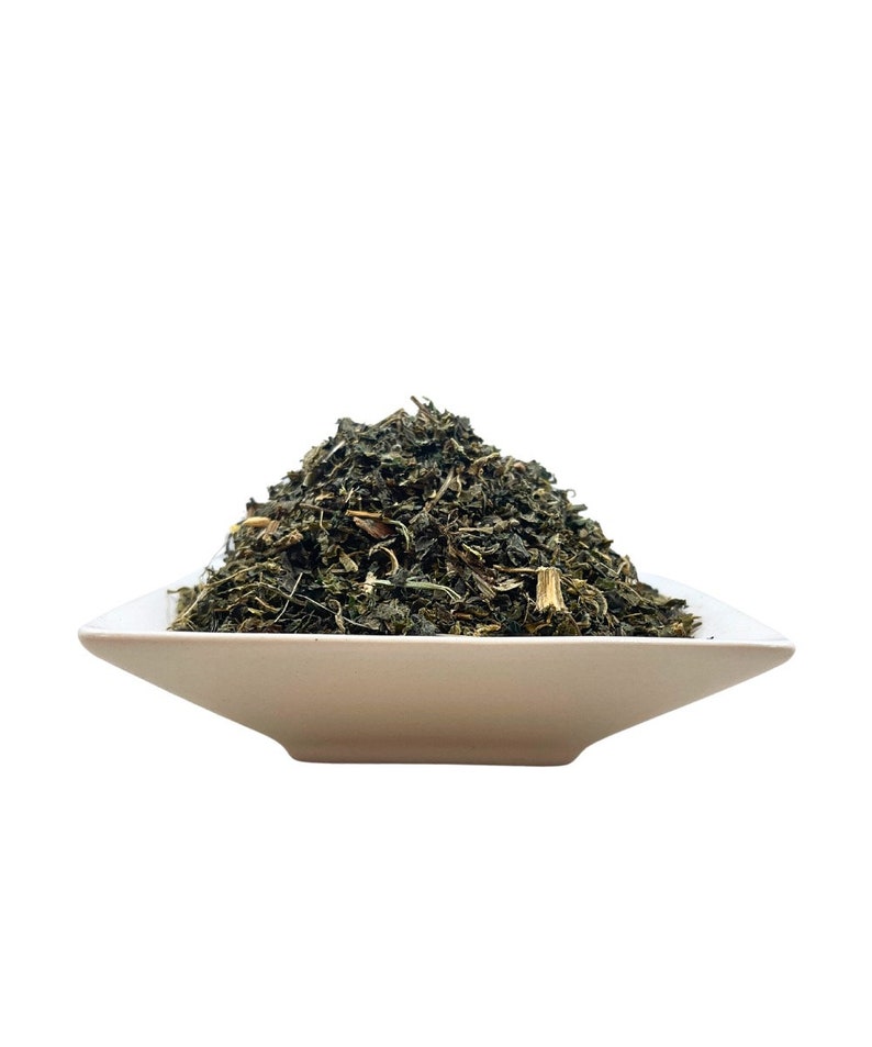 May include: A mound of dried, dark green herbal leaves and small stems piled high in a square, off-white ceramic bowl. The texture of the dried plant matter is coarse and varied, featuring shades of deep forest green and lighter brown accents.