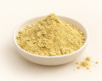 May include: A small white ceramic bowl filled with a mound of fine, pale yellow powder. The texture of the powder appears dry and slightly granular, with a few small clumps visible throughout the pile. A small amount of powder has spilled.